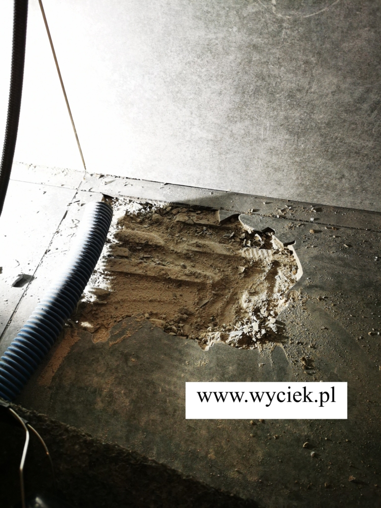 Punktowa odkrywka w posadzce przy ścianie łazienki i wąż odkurzacza obok usuniętych płytek.