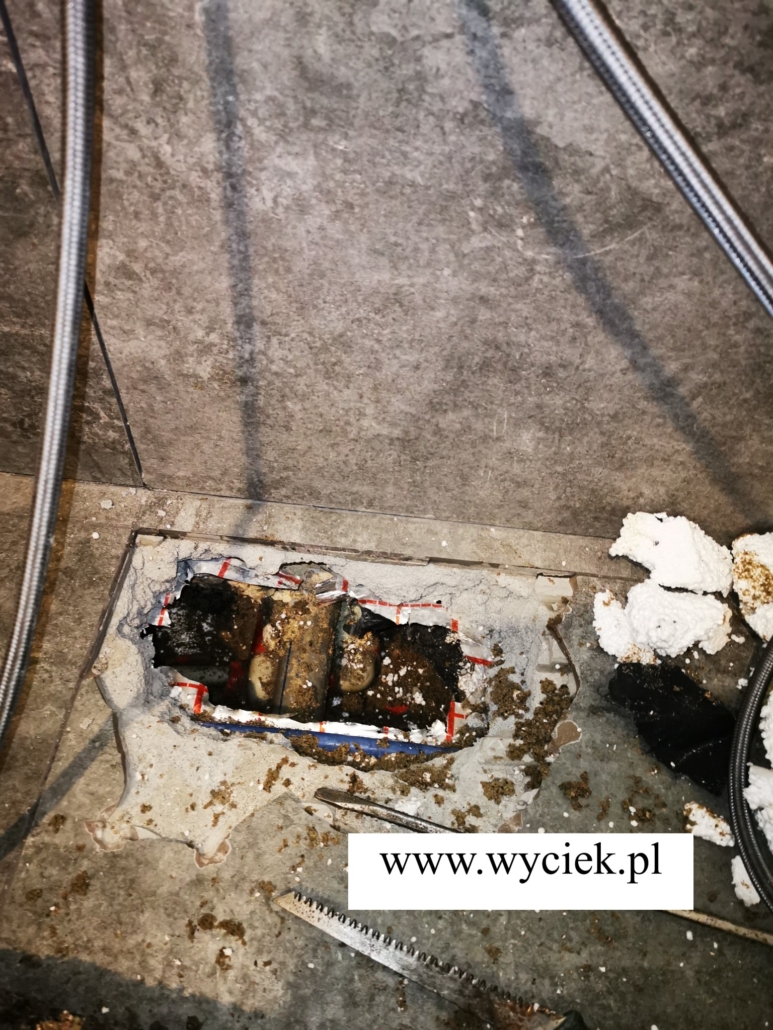 Rury i warstwy izolacji odsłonięte w punktowej odkrywce pod posadzką przy ścianie.